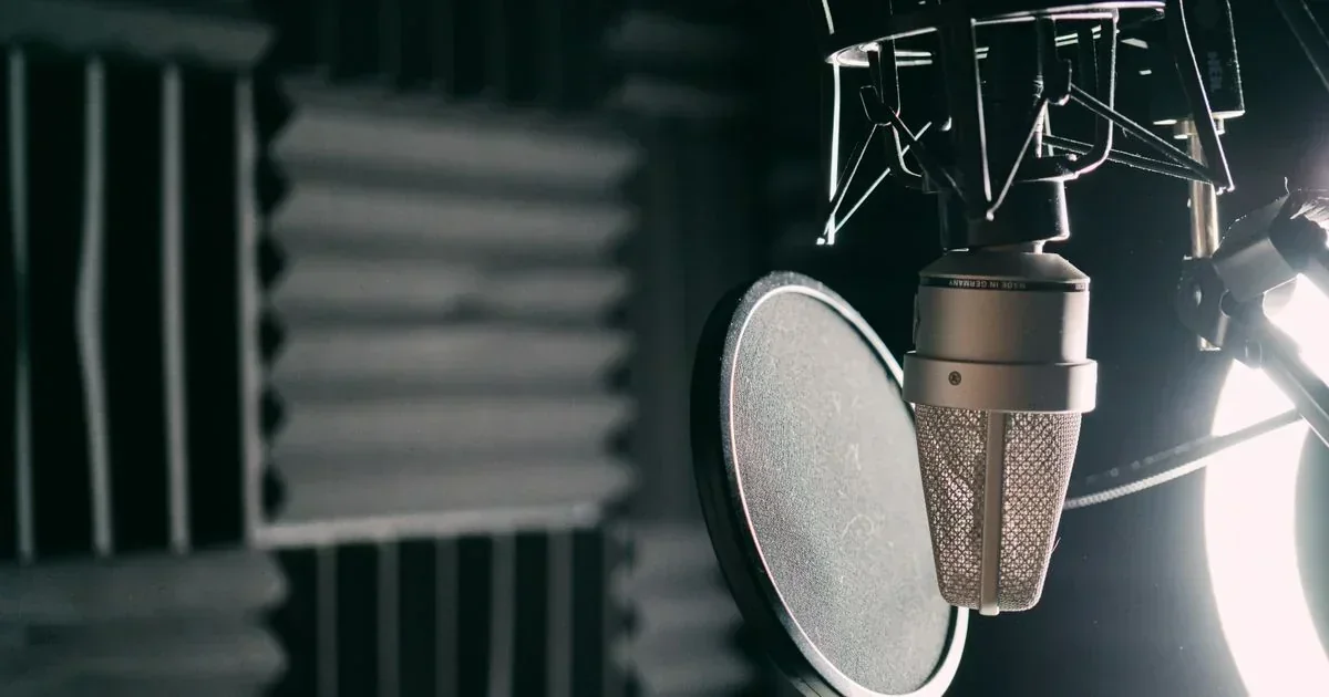 A silver condenser microphone with a pop filter in a recording studio, with acoustic foam panels and a bright light in the background.