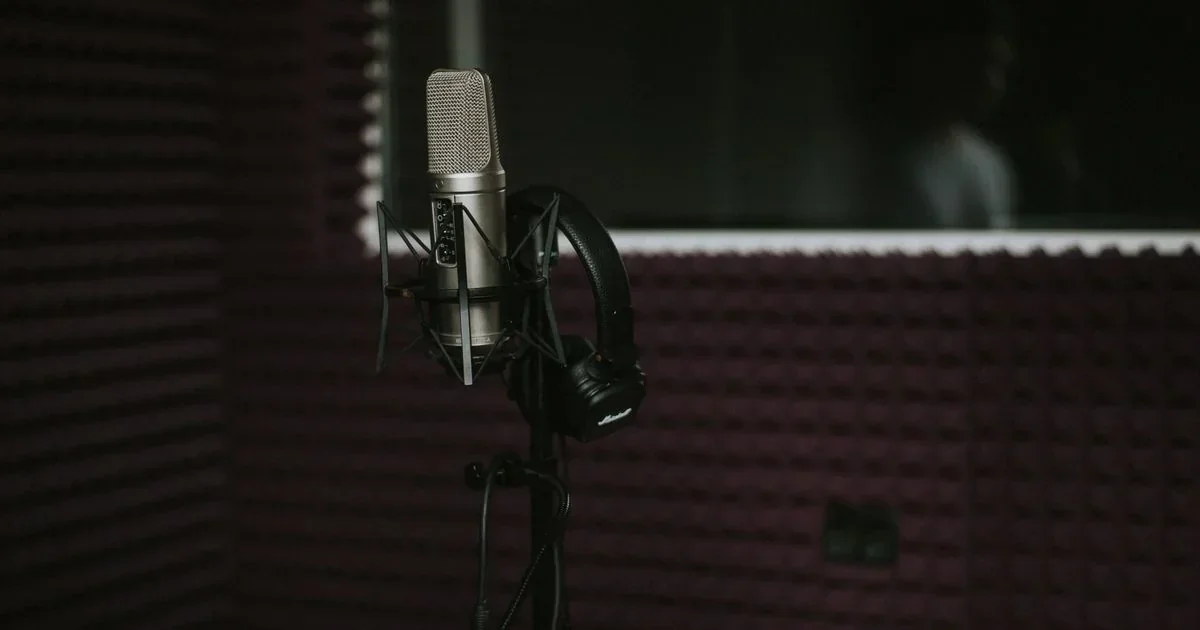 A silver studio condenser microphone in a shock mount with black headphones resting on a stand inside a dimly lit podcast recording booth with acoustic foam panels.