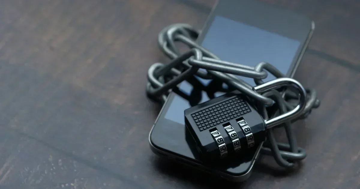 A black smartphone wrapped in a metal chain secured by a combination padlock on a dark wooden surface.