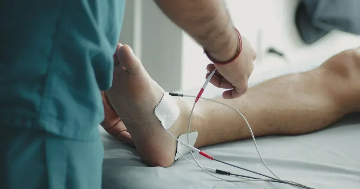 A clinician in teal scrubs attaches electrode wires to a patient's bare foot during a physical therapy session.