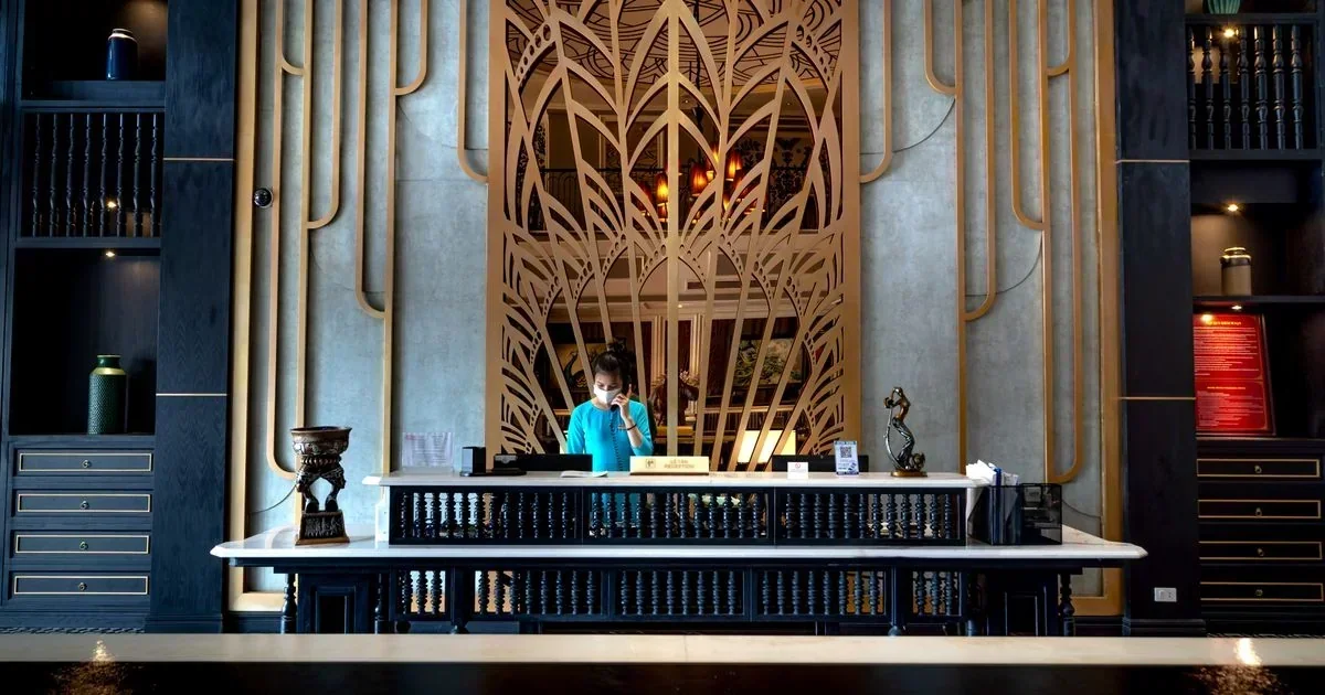 A hotel receptionist in a teal uniform speaks on the phone behind an ornate white and black front desk with gold Art Deco screens.