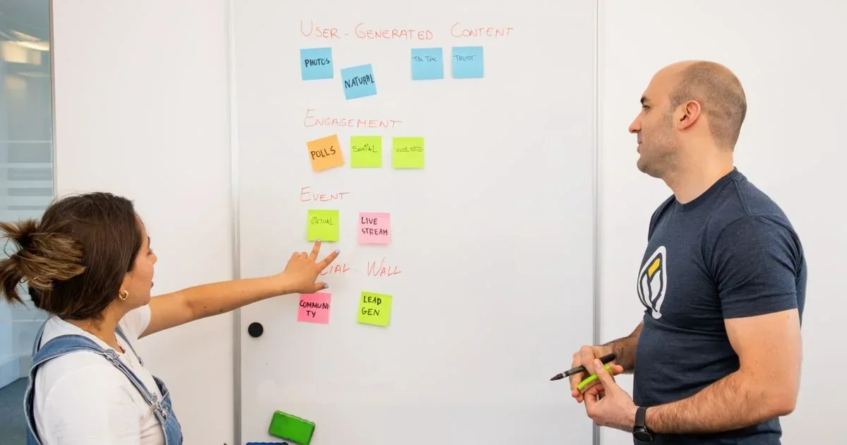 Woman pointing at colorful sticky notes on a whiteboard labeled User-Generated Content, Engagement, and Event while a man holding a pen watches during a brainstorming session.
