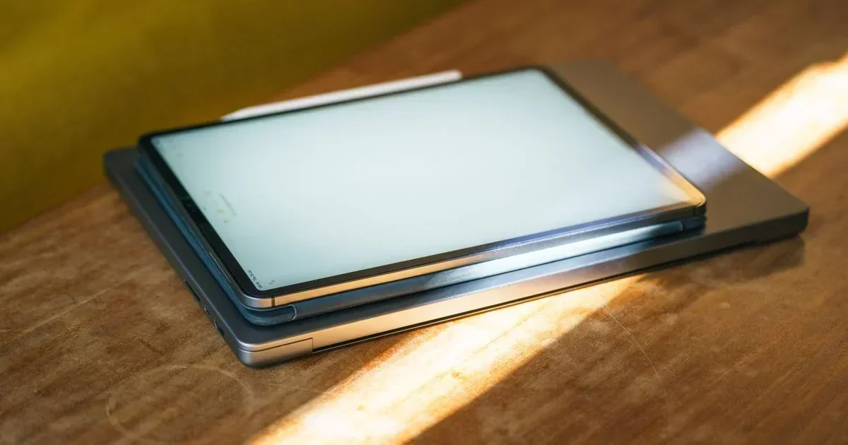 A tablet with a stylus resting on a closed laptop on a wooden desk, illuminated by warm sunlight.