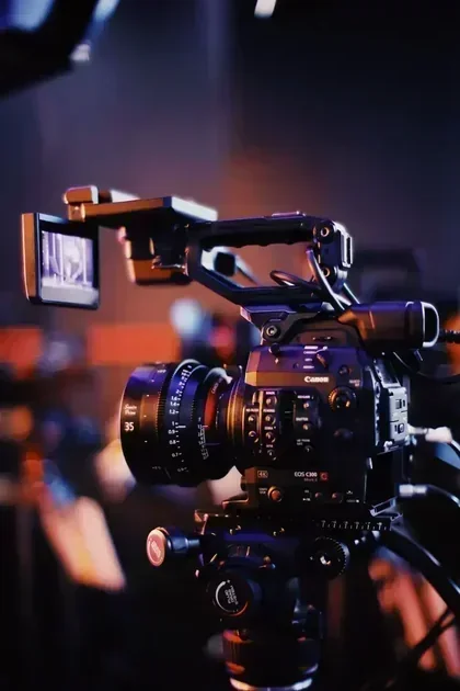 A Canon EOS C100 cinema camera with a side-mounted monitor and cine lens sits on a tripod in a dimly lit studio.