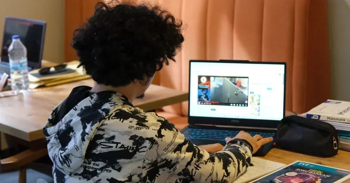 Student in a black-and-white graphic hoodie using an MSI laptop showing a video lesson at a wooden desk with textbooks and a water bottle.