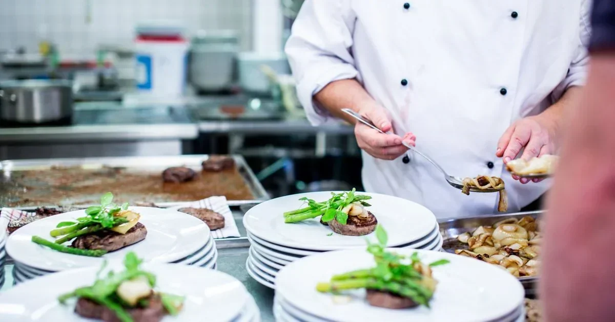 Chef in white uniform plating sliced steak with green garnish and onions onto white plates in a commercial kitchen.