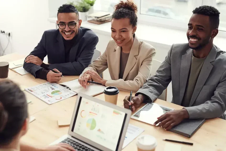Three smiling professionals in business attire review printed analytics charts and a laptop displaying a pie chart during a collaborative meeting at a wooden table.