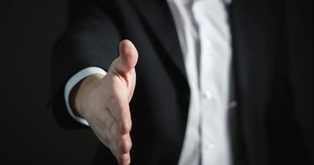 Close-up of a businessman in a suit extending an open hand for a handshake against a dark background.