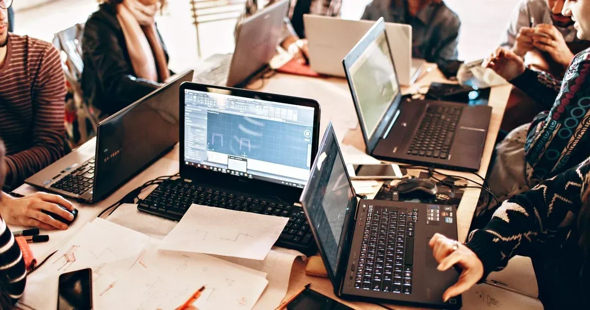 A team collaborates around a table covered in open laptops, wireframe sketches, and a smartphone, with a code editor visible on one screen.