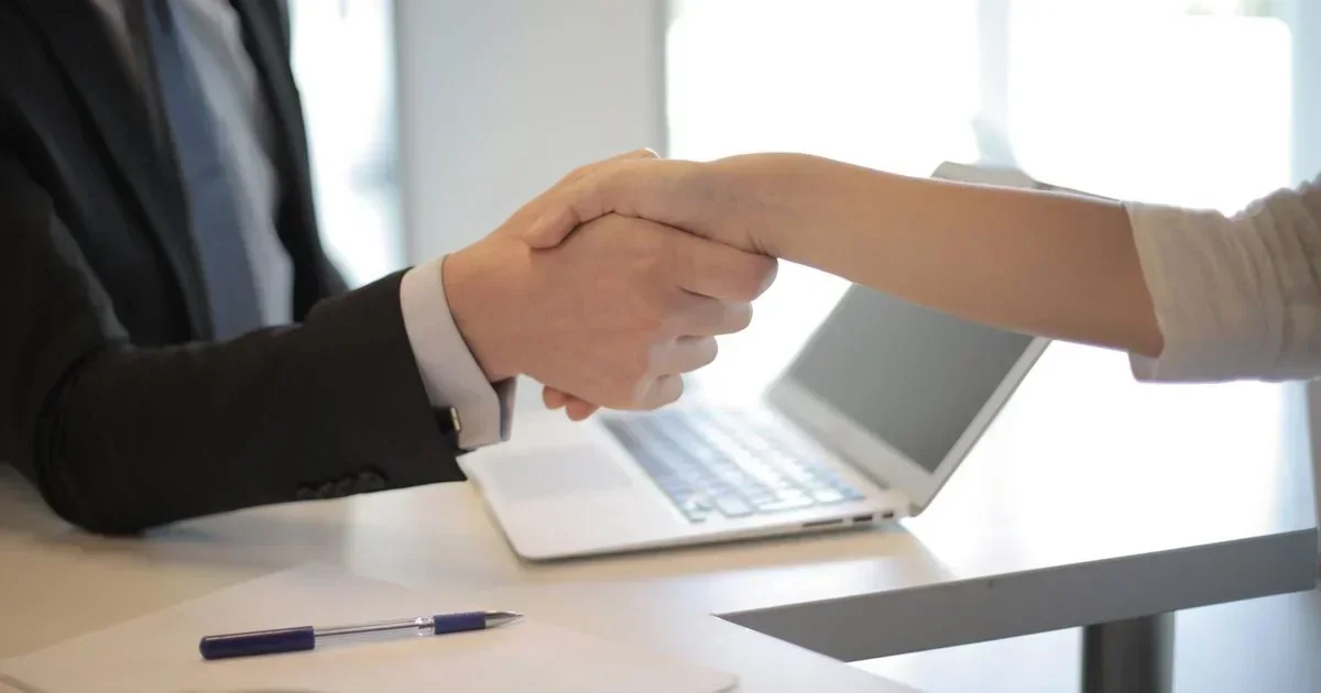 Two people in business attire shaking hands across a white desk with an open laptop, notepad, and pen during an office meeting.