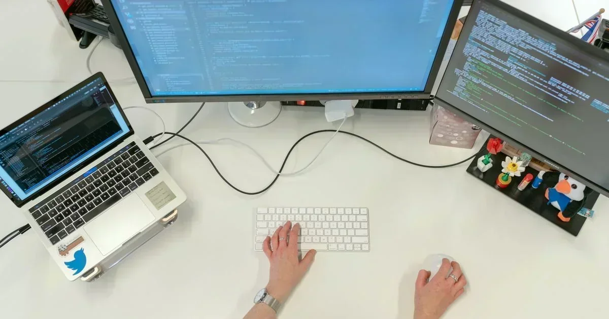 Overhead view of hands typing on a white keyboard and using a mouse at a desk with a laptop and two monitors displaying code.