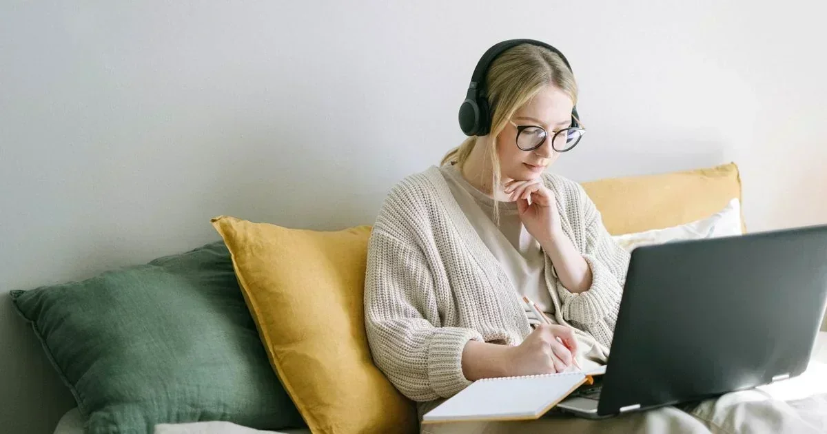 Blonde woman in headphones and glasses writing in a notebook while watching a laptop, sitting against green and yellow pillows.