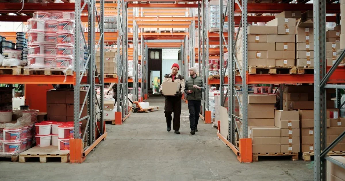 Two warehouse workers carrying a box walk down an aisle lined with tall orange pallet racks stacked with shrink-wrapped goods and cardboard boxes.