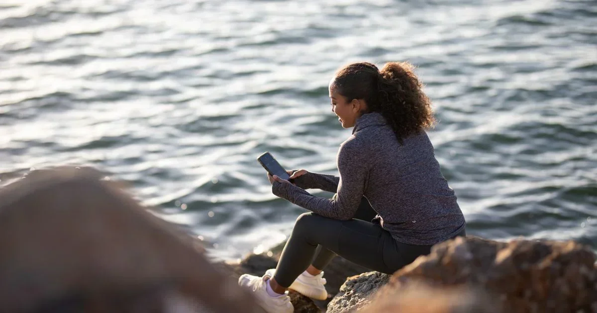Smiling woman in athletic wear sitting on coastal rocks using a smartphone with the ocean behind her.