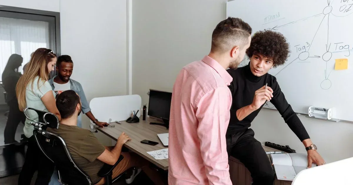 A team of five colleagues collaborates in an office, with two men discussing a workflow diagram on a whiteboard while others work at a desk.
