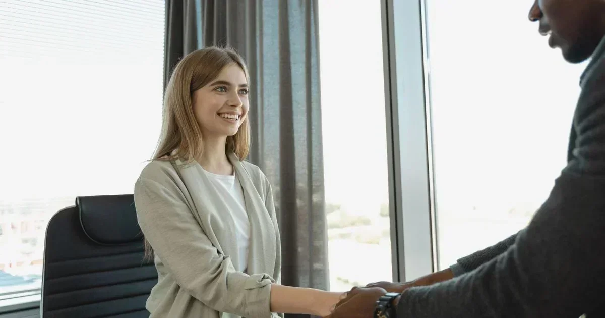 Smiling blonde woman in a beige blazer shaking hands with a man in an office, likely concluding a job interview or HR meeting.