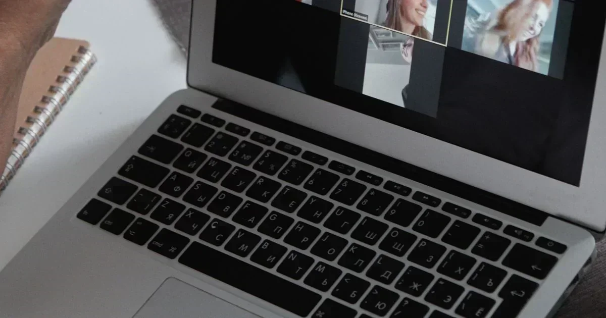 A close-up of a laptop with a Cyrillic keyboard displaying a video call with two women, next to a spiral notebook.