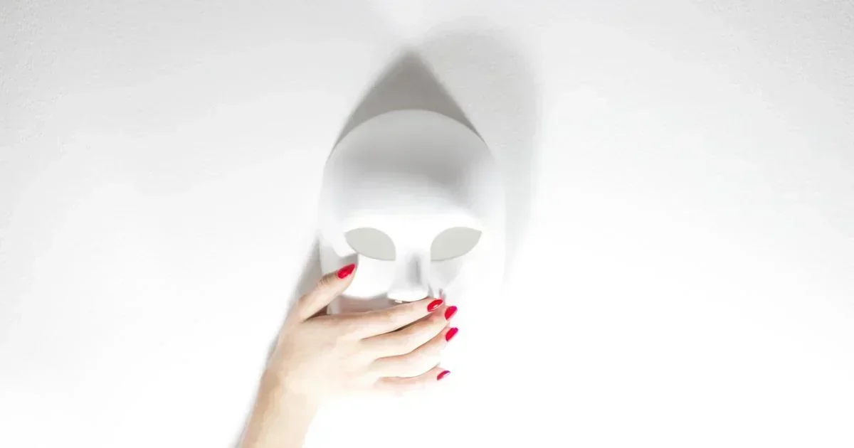 A hand with red nail polish holds a blank white face mask against a white background, casting a shadow.