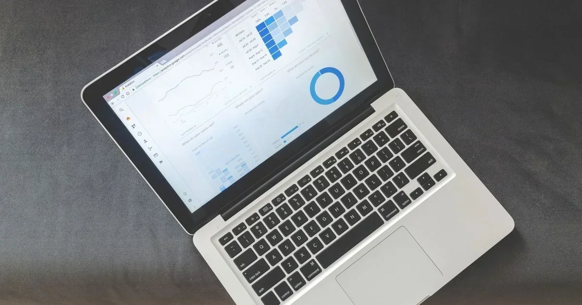 An open silver laptop on a gray surface displays a Google Analytics dashboard with a line graph, blue bar chart, and donut chart.