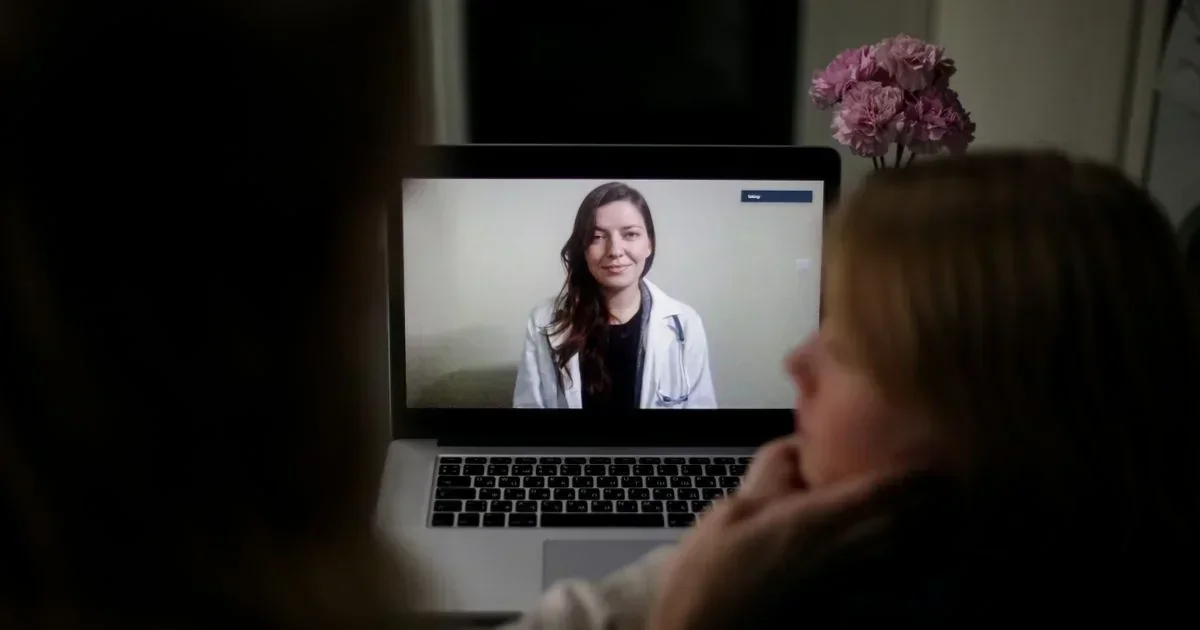 A patient views a female doctor in a white coat wearing a stethoscope during a telemedicine video call on a laptop screen.