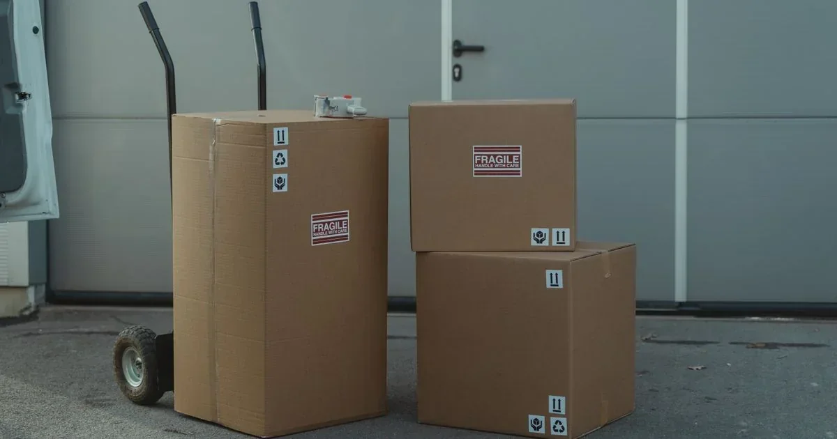 Three cardboard shipping boxes with fragile labels sit on a loading dock, one stacked on a hand truck, outside a gray warehouse door.