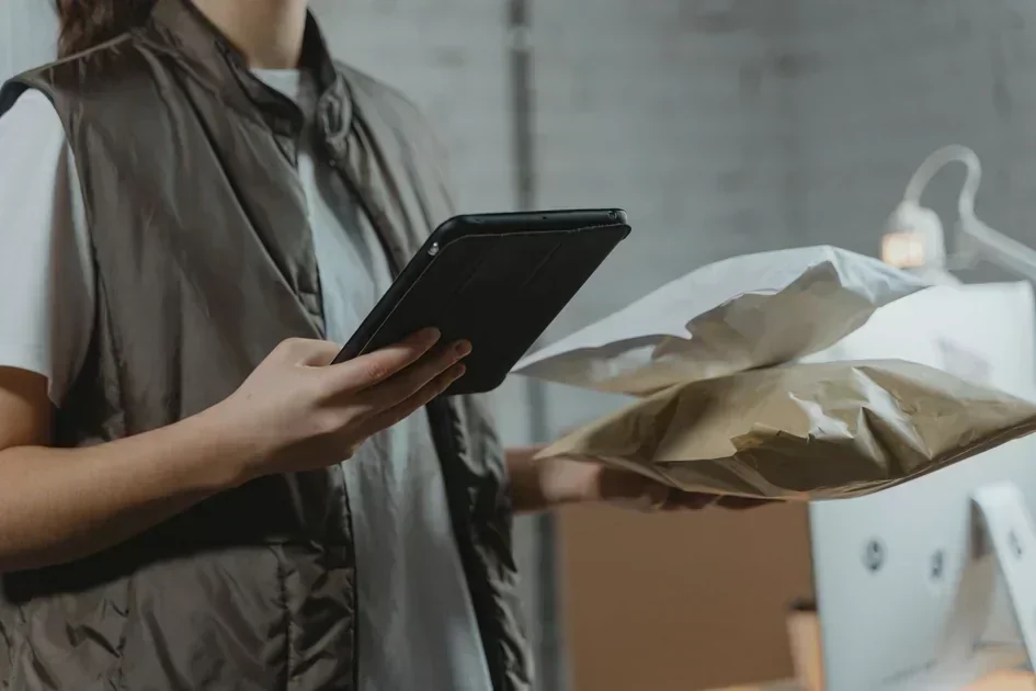 Person in a vest holding a tablet and two padded shipping envelopes beside a computer.