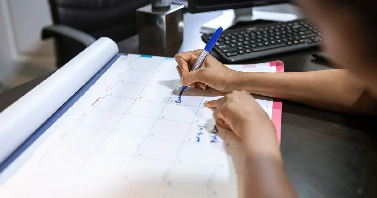 Person writing a meeting note in blue ink on a large paper monthly calendar at a desk with a keyboard.
