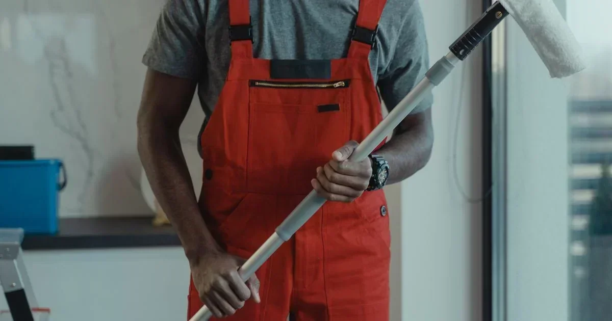 A cleaning worker in red overalls holds a long-handled duster while standing in a bright room with a blue bucket and ladder.