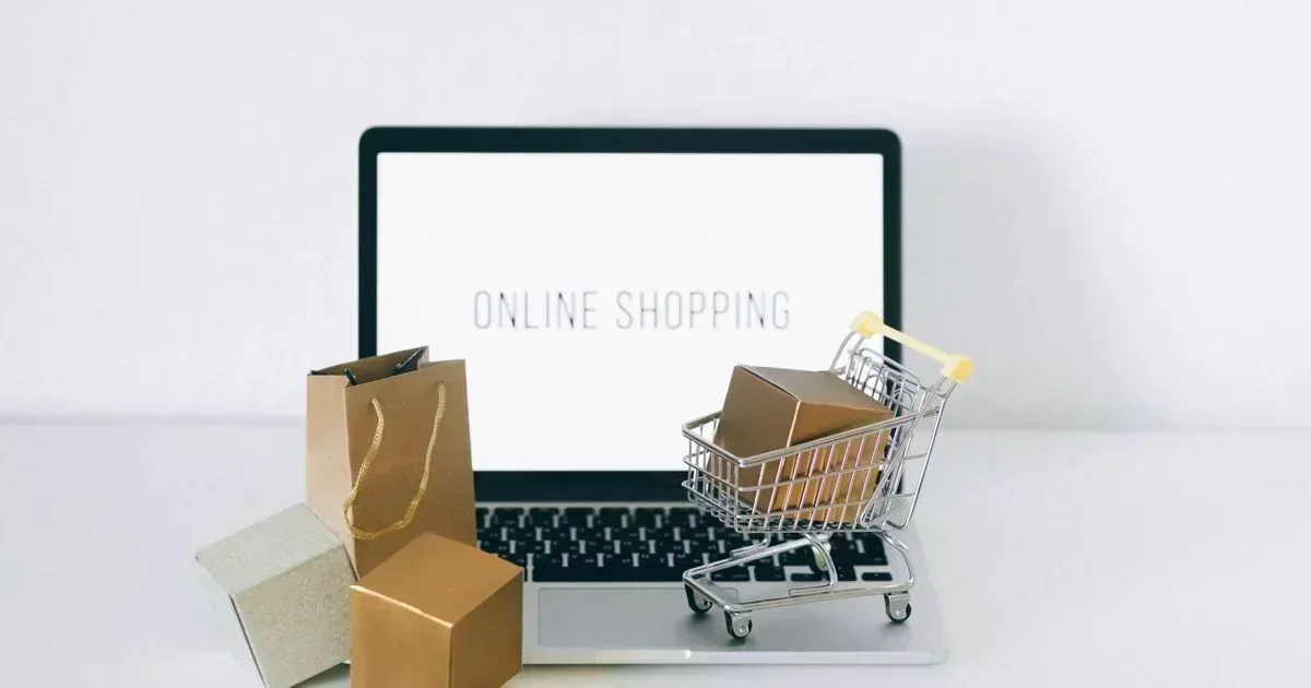 A laptop displaying "ONLINE SHOPPING" on its screen sits behind a miniature shopping cart holding a small box, a brown paper bag, and two gift boxes on a white surface.