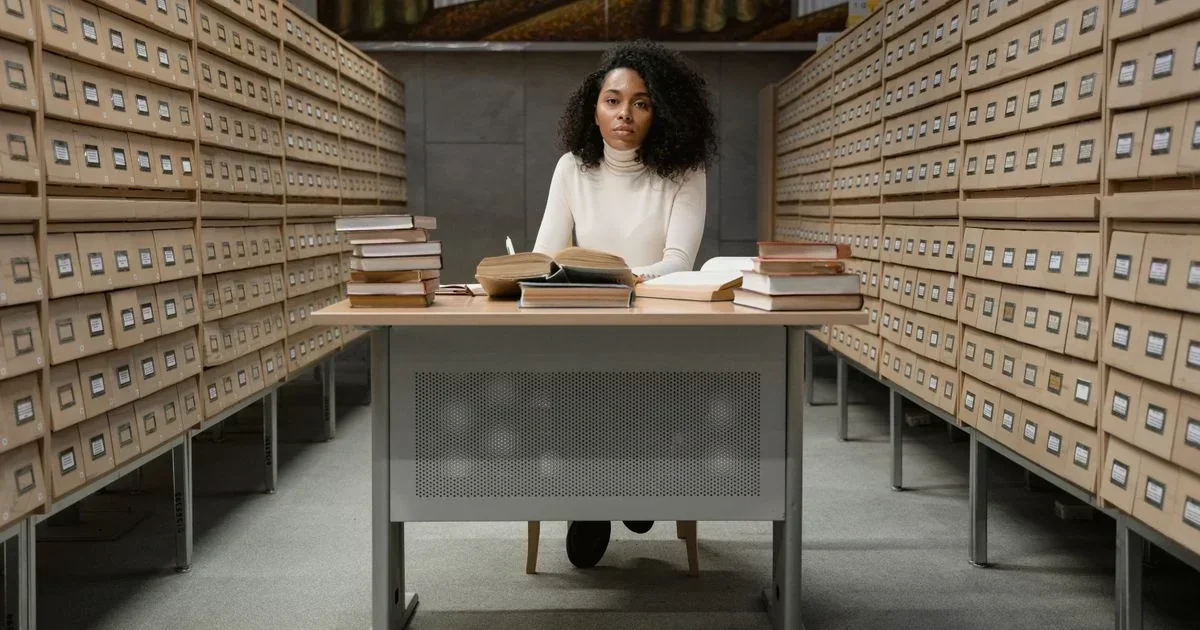Woman seated at a desk with open books between towering rows of archive drawers in a dim research room.