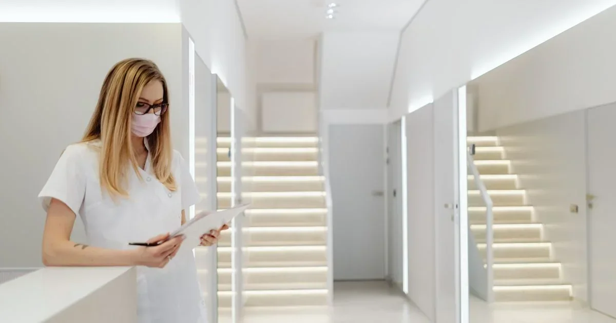 Masked female healthcare worker in white scrubs writing on a clipboard in a bright, modern clinic hallway with illuminated stairs.