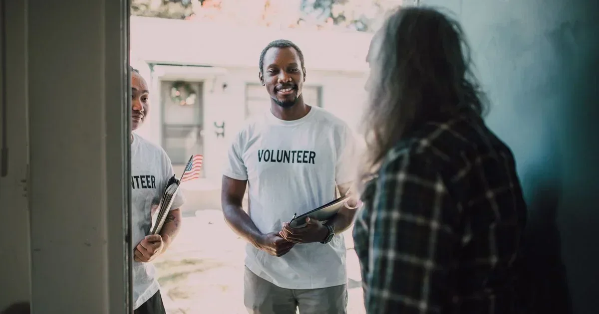 Two smiling volunteers in white Volunteer t-shirts holding clipboards stand at a doorway speaking with a resident inside a home.