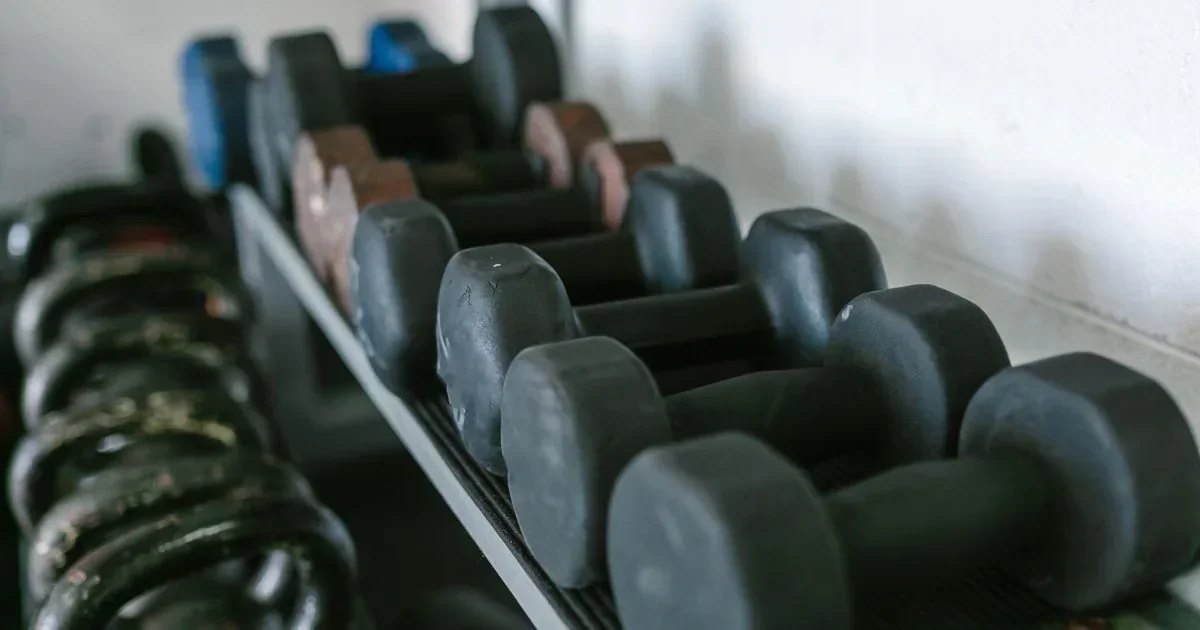 Rows of black, blue, and brown hex dumbbells on a rack beside stacked weight plates in a gym.