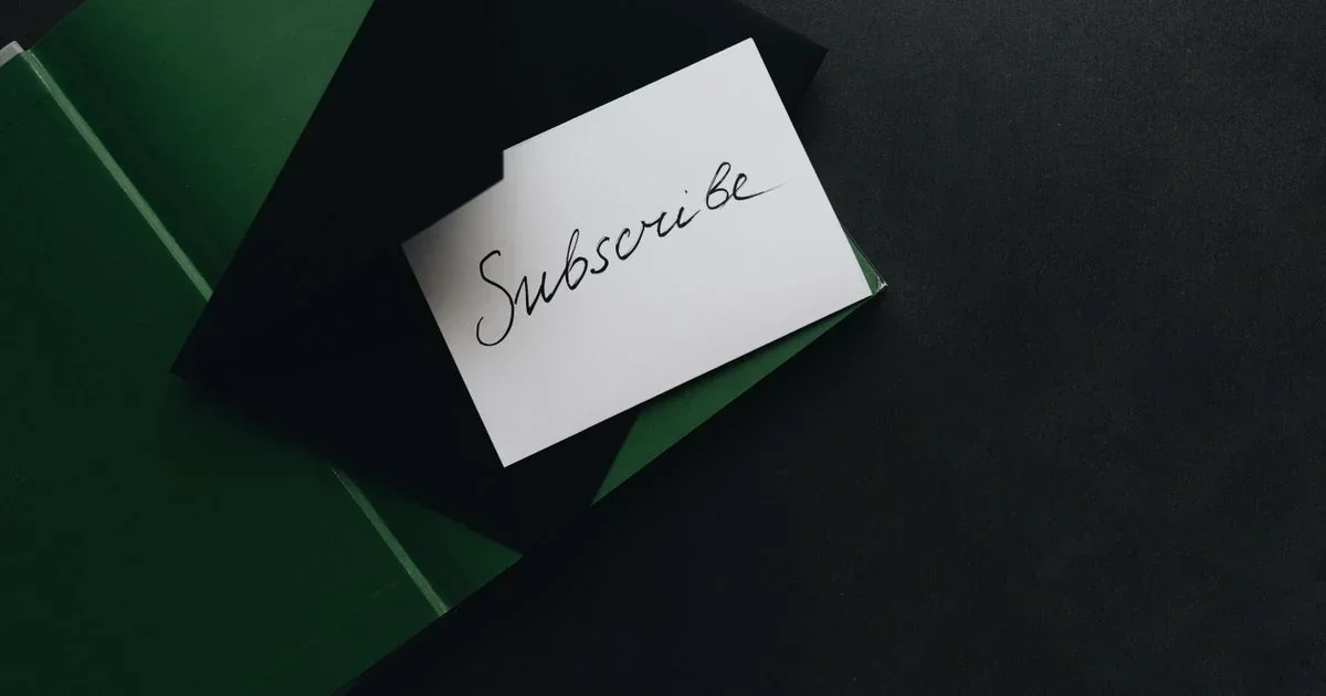 A white card with the handwritten word Subscribe in cursive rests on dark green folders against a black background.