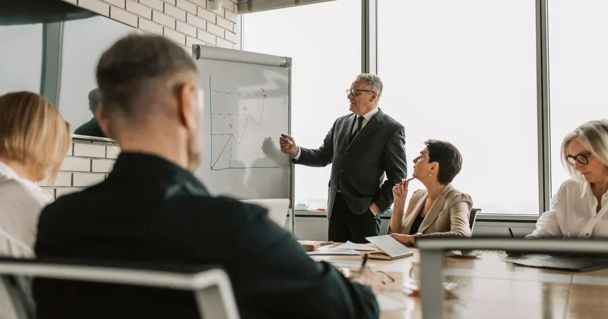 A businessman presents a line graph on a whiteboard to seated colleagues in a bright conference room.