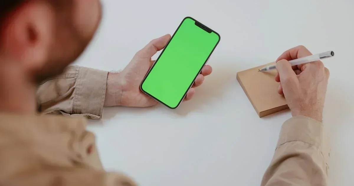 Over-the-shoulder view of a man in a tan shirt holding a smartphone with a green screen in one hand and a pen over a small notepad in the other.