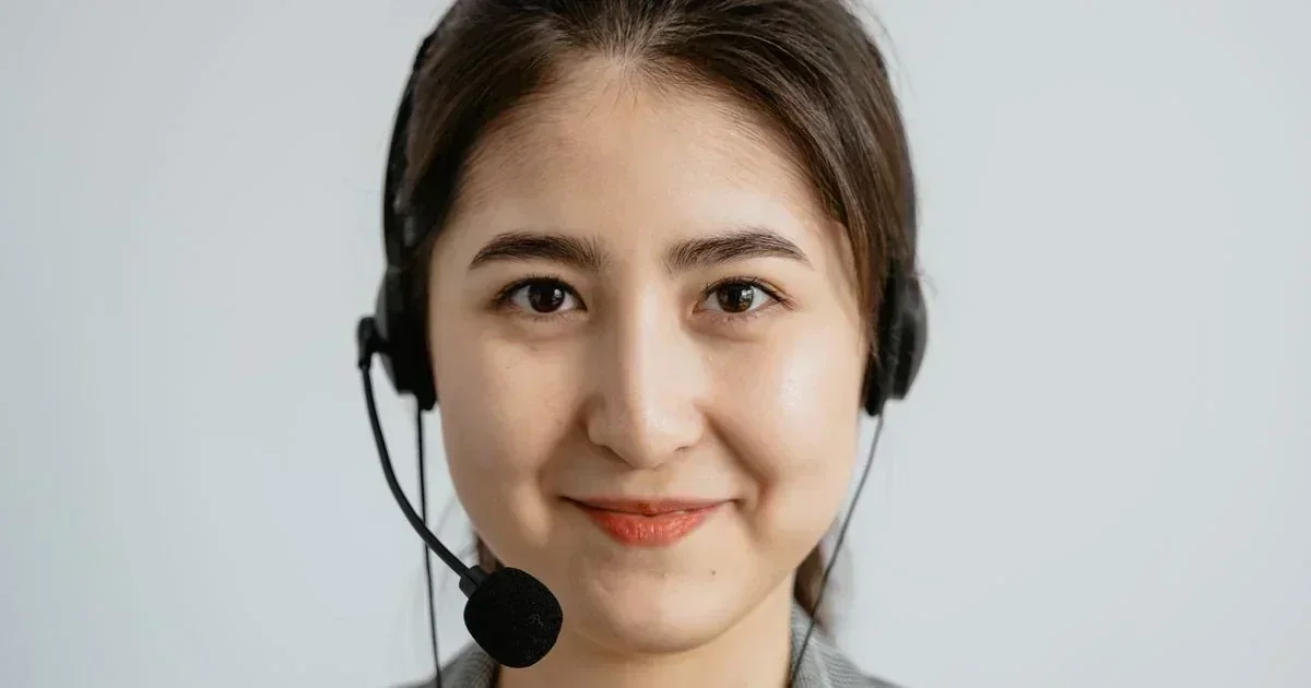 Smiling female customer service agent wearing a black headset with microphone against a plain light gray background.