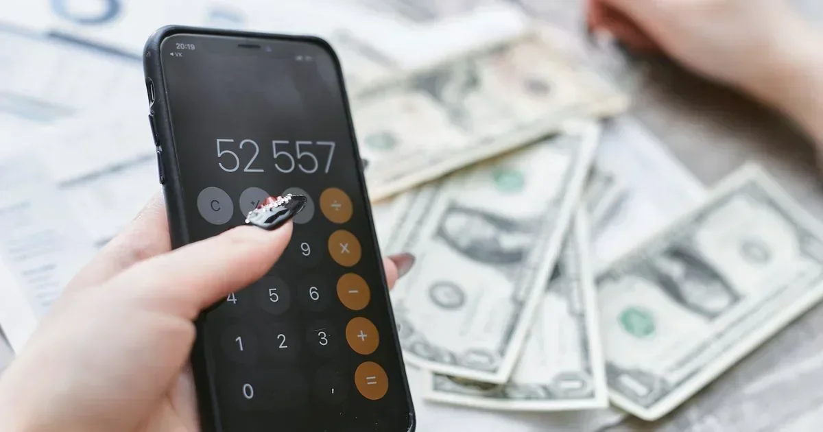 A hand taps a smartphone calculator showing 52,557 above scattered US hundred-dollar bills and financial paperwork.