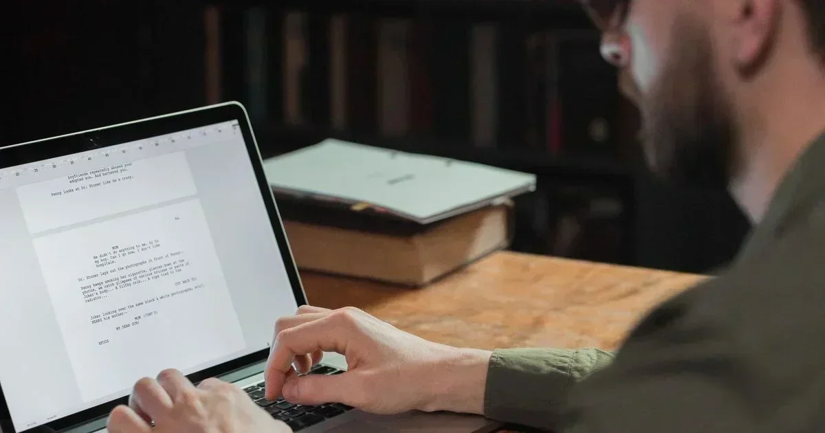 A bearded man types on a laptop displaying a document, seated at a wooden desk with a book in the background.