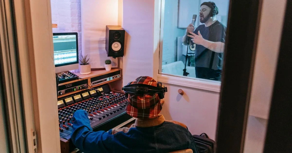 Music producer in plaid hat at a mixing console with waveform on screen, watching a singer record vocals through a studio window.