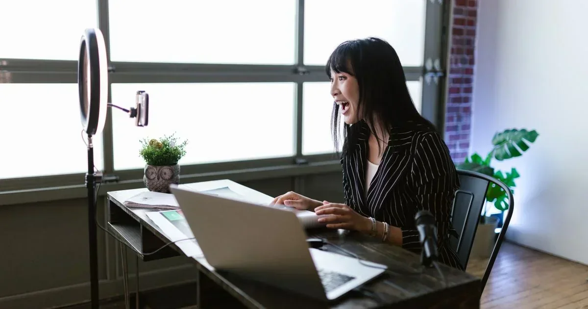 Woman in striped blazer laughing while recording video at a desk with a ring light, phone mount, laptop, and microphone.