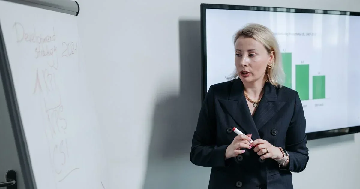 Blonde businesswoman in a black blazer holding a marker beside a whiteboard and a screen displaying a green bar chart.