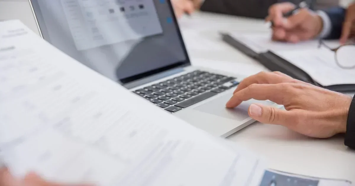 A close-up of hands using a laptop touchpad beside printed documents on a white desk during a business meeting.
