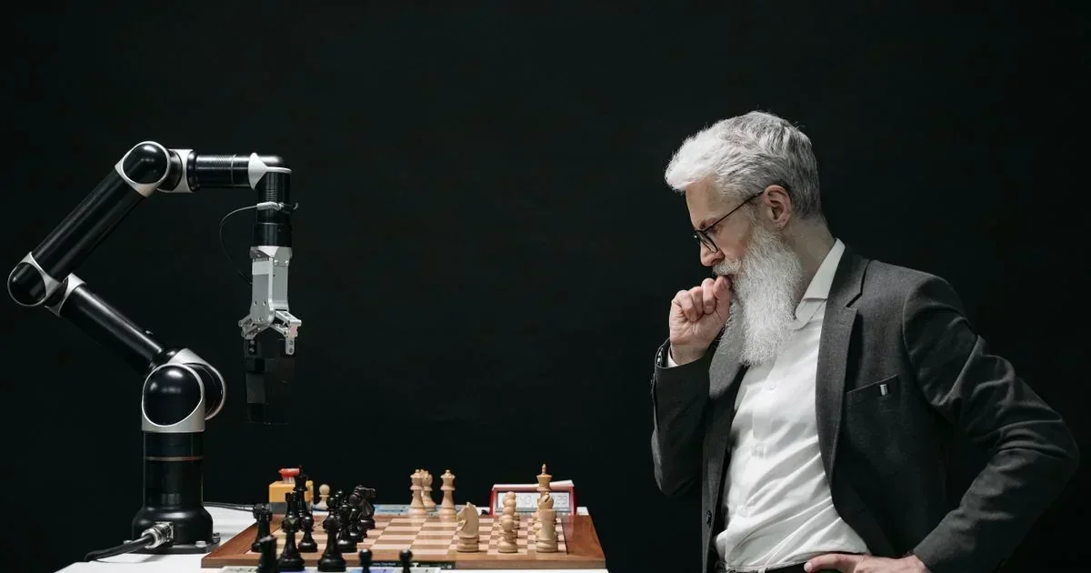 An older man with a white beard contemplates a chessboard opposite a robotic arm under a black background.
