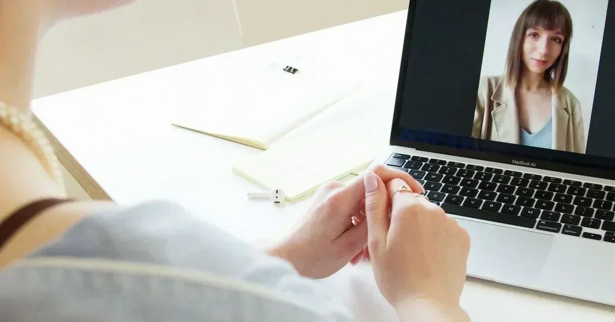Over-the-shoulder view of a person with clasped hands watching a woman on a MacBook Air during a video call, with a notebook and earbud on a white desk.