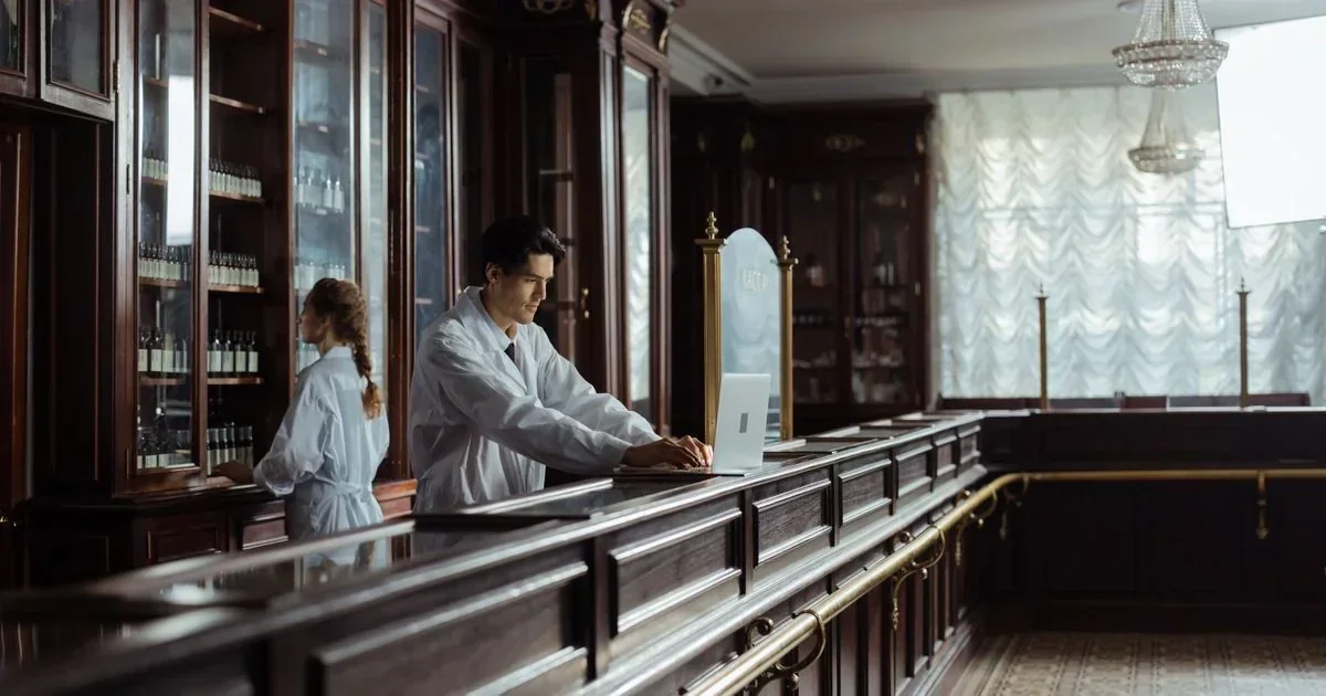 Pharmacists in white coats working behind a dark wood counter with glass cabinets of medicine bottles, one using a laptop.