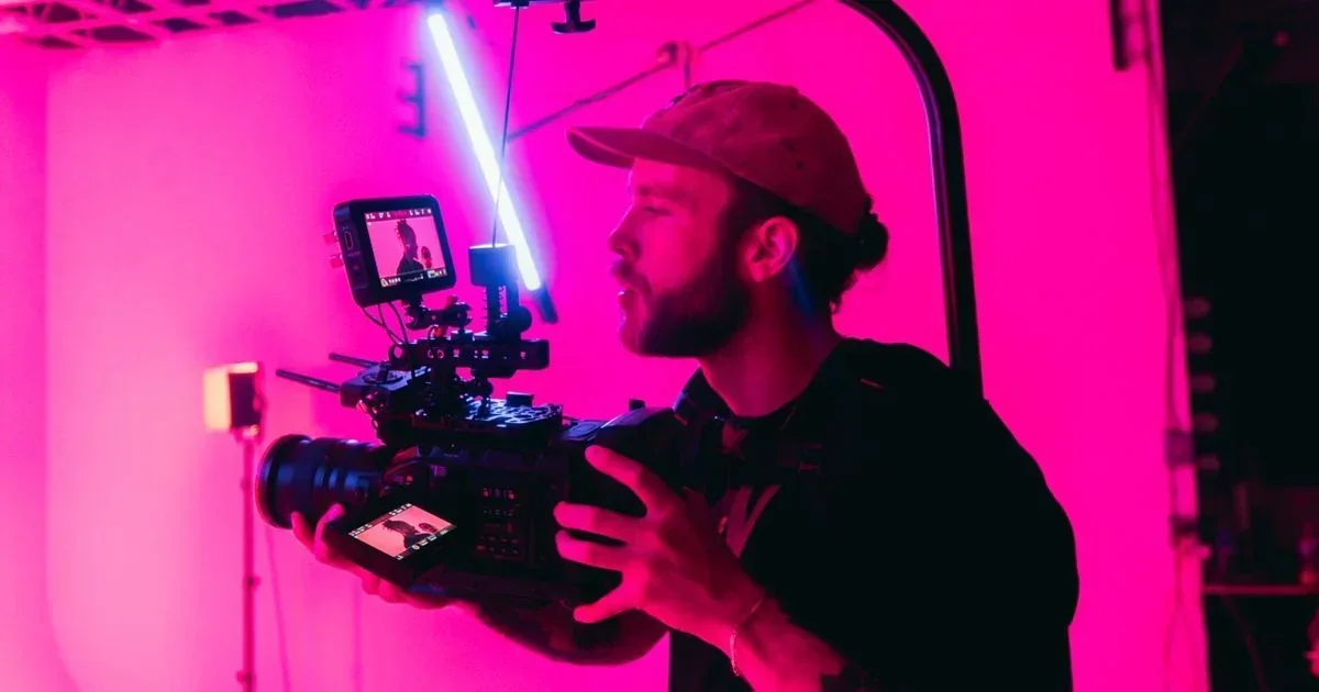Bearded videographer in a cap operates a cinema camera with a monitor under bright pink studio lighting.