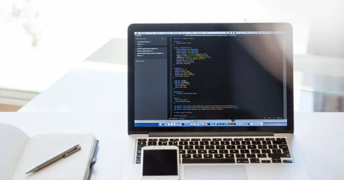 Laptop displaying a Python code editor on a white desk beside a notebook, pen, and smartphone.