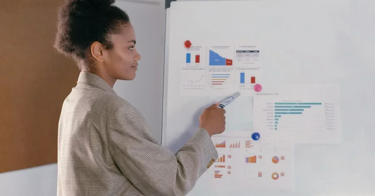 Woman in a plaid blazer pointing a marker at printed analytics charts pinned to a whiteboard during a marketing strategy review.