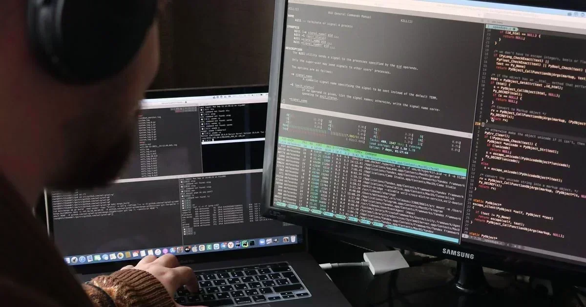A person wearing headphones types on a laptop while viewing Python code and terminal output across a laptop and Samsung monitor.